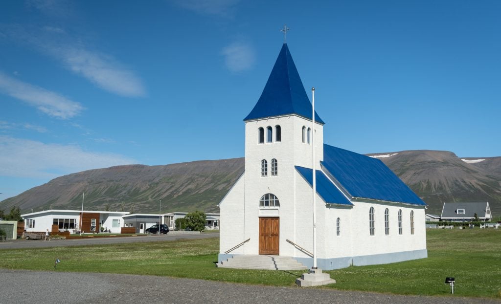 A small white church with a royal blue steeple and roof, in front of small cottages.