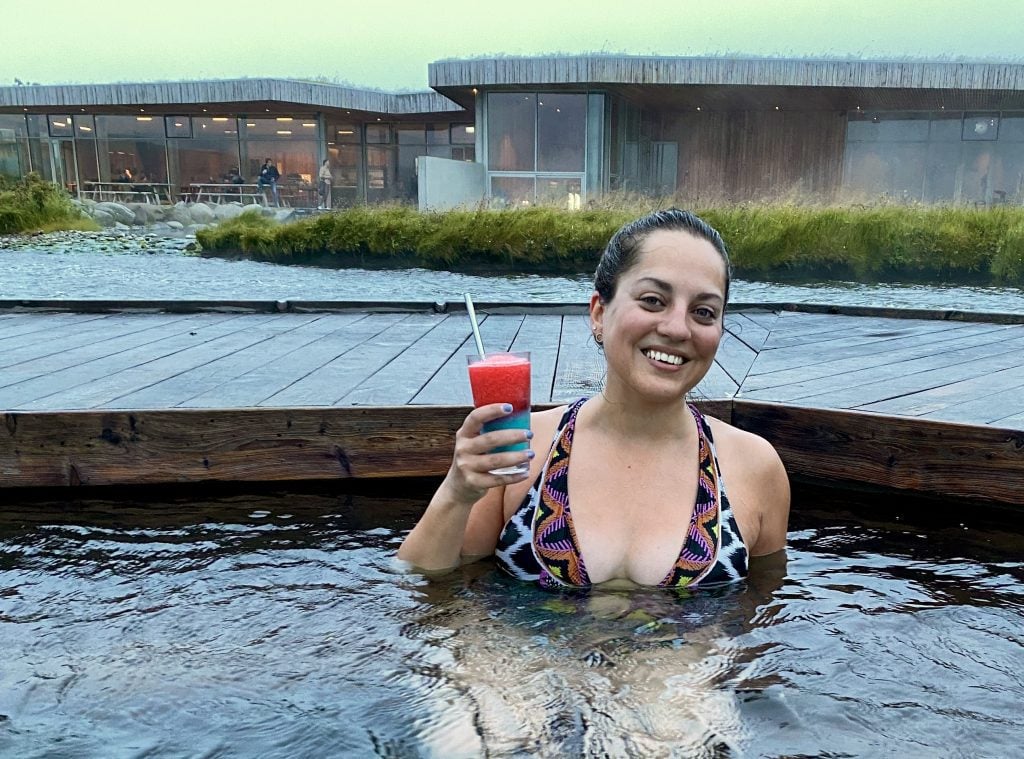Kate smiling and holding up a bright red and blue slushy while sitting in a hot pool.