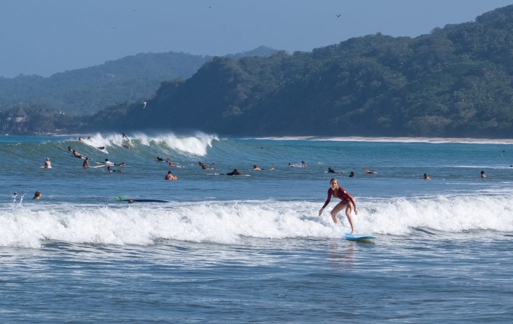 A woman surfer hanging 10 over a wave, a few dozen surfers on their bellies on their boards behind her.