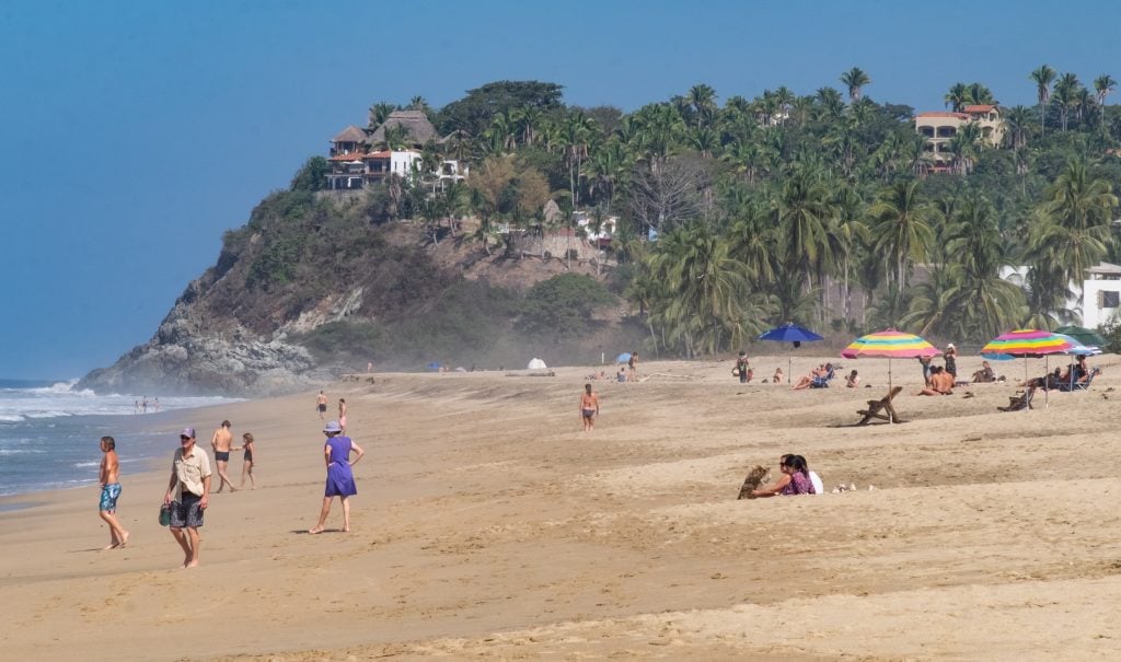 People and dogs hanging out on a long sandy beach, a jungle-topped mountain in the background.