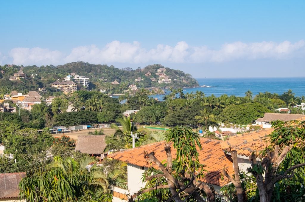 A view of hills topped with palm trees and orange-roofed villas leading to the bright blue ocean.