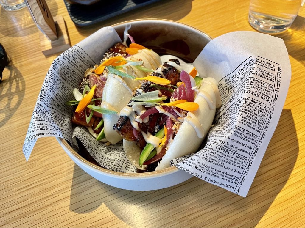 Two decorated bao buns filled with pork belly wrapped in fake newsprint in a bowl.