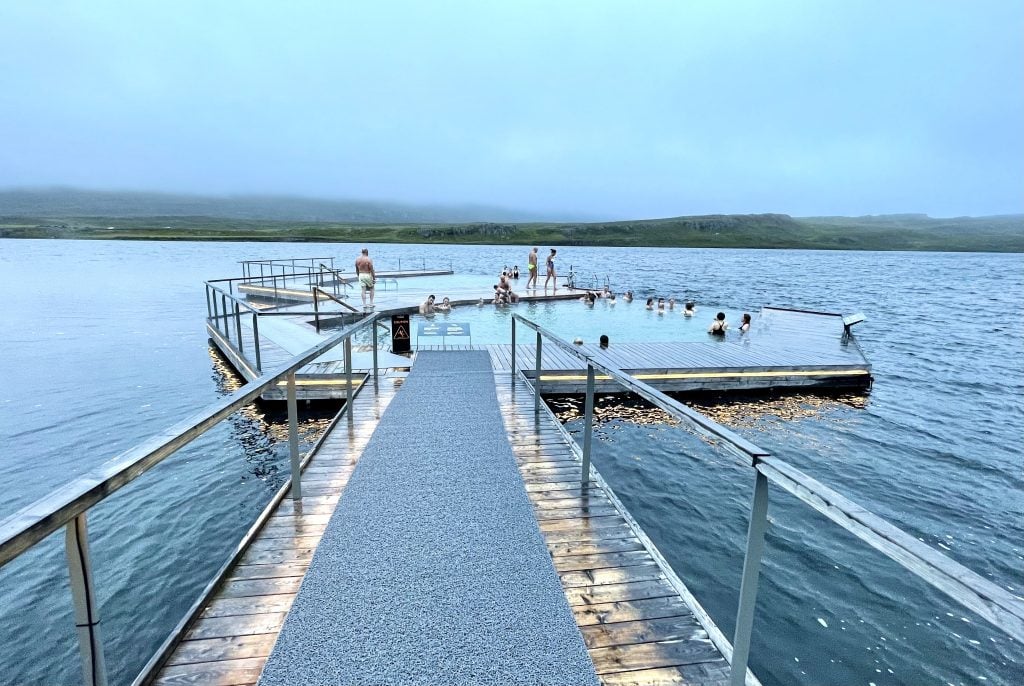 A pathway leading across the lake to two floating pools filled with about 30 people.
