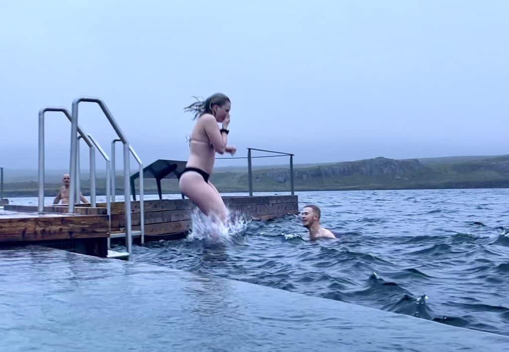 A girl in a bikini jumping off the deck into the cold lake, holding her nose.