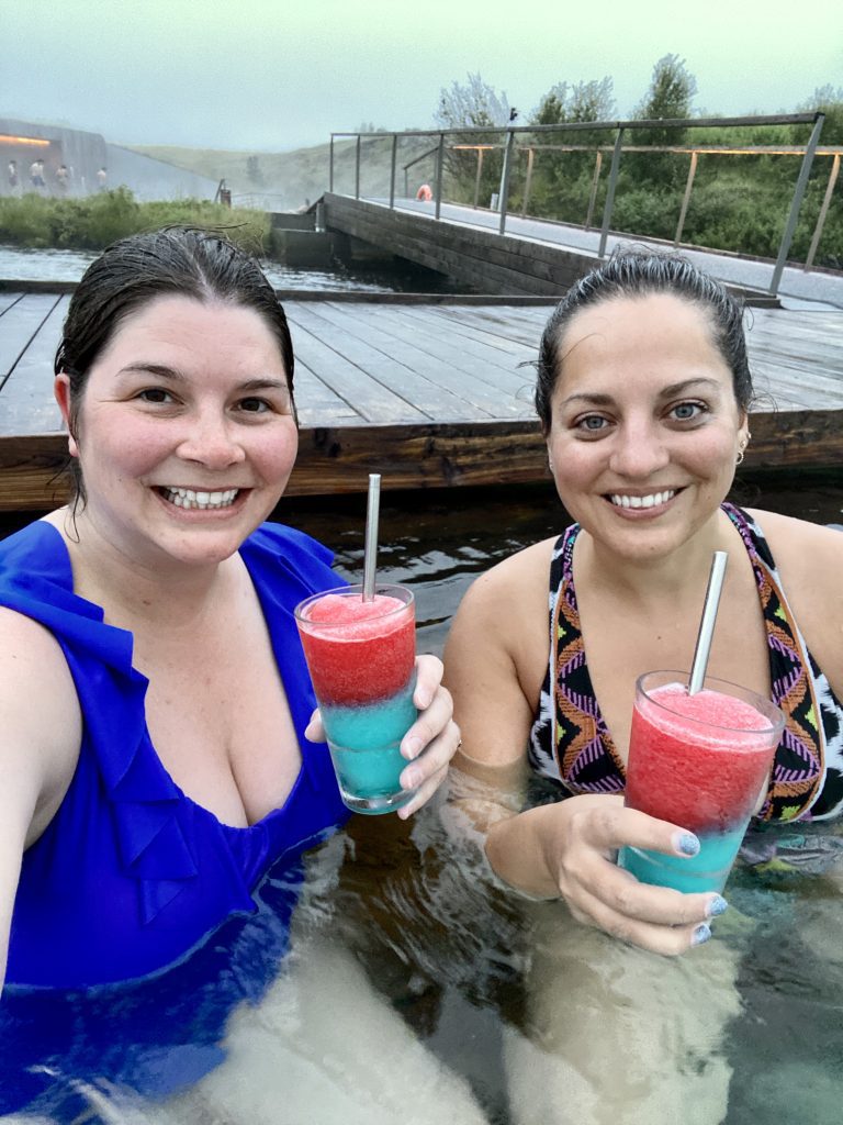 Kate and Amanda taking a selfie and smiling, in the pool wearing bathing suits, each with a bright red and blue slushy in hand.