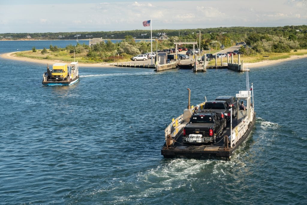 A teeny tiny ferry that can only fit three cars and sits flat on the water, heading for a grassy green island.