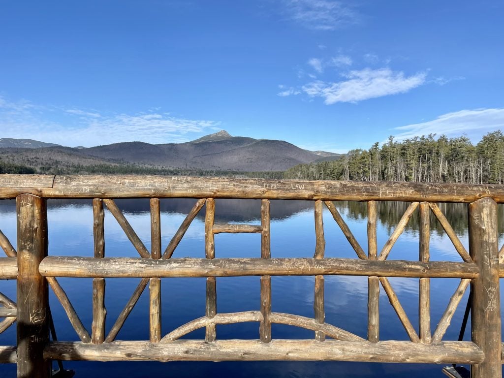 A still blue lake with mountains in the background and a wooden fence in front of it.