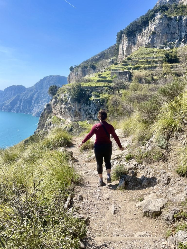 Kate hiking down a dirt path set against tall bright green cliffs, the bright blue sea below.