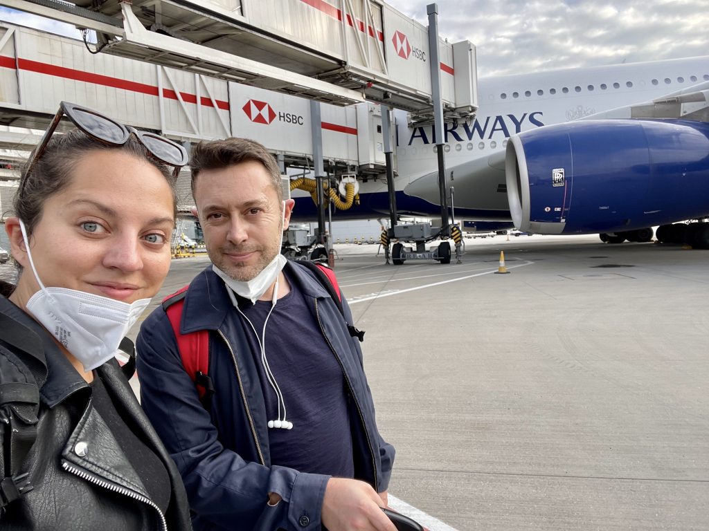 Kate and Charlie take a selfie in front of a British Airways plane, their masks around their chins.