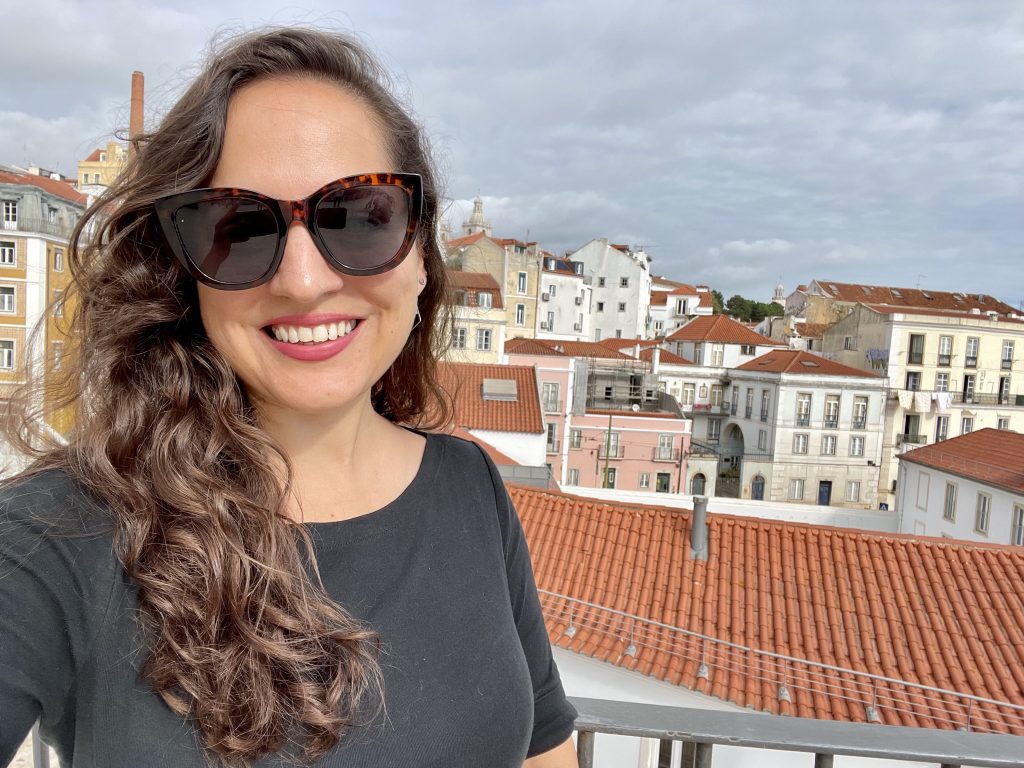 Kate takes a smiling selfie with sunglasses and long curly brown hair, the orange roofs and white buildings of hilly Lisbon behind her.