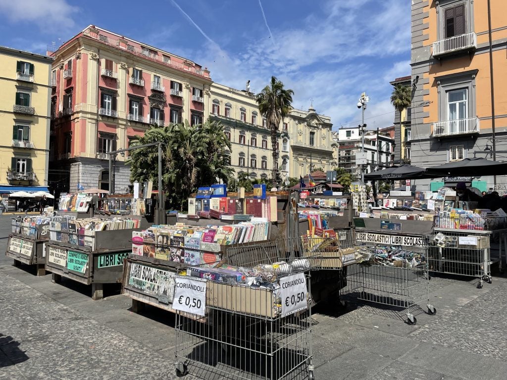 A street market on a pizza surrounded by pink and yellow buildings and palm trees. Multiple tables selling piles of books.