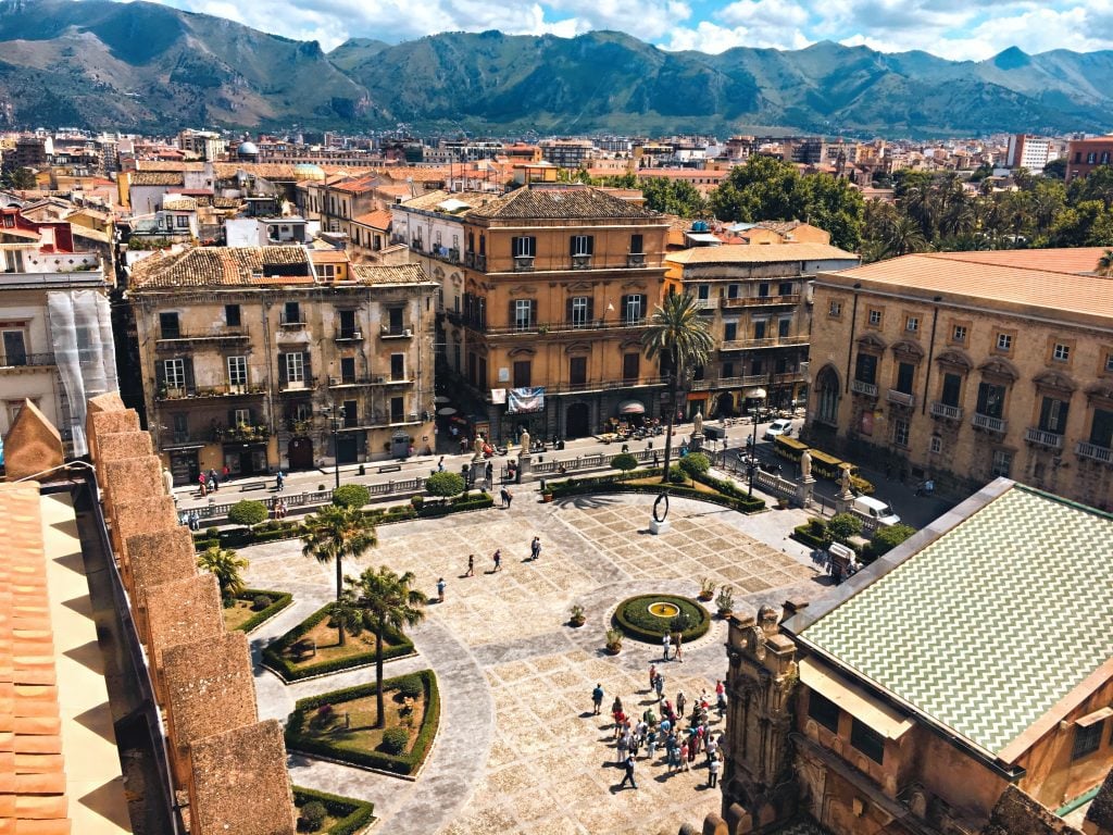 A large piazza surrounded by sand-colored buildings in Palermo, mountains in the distance.