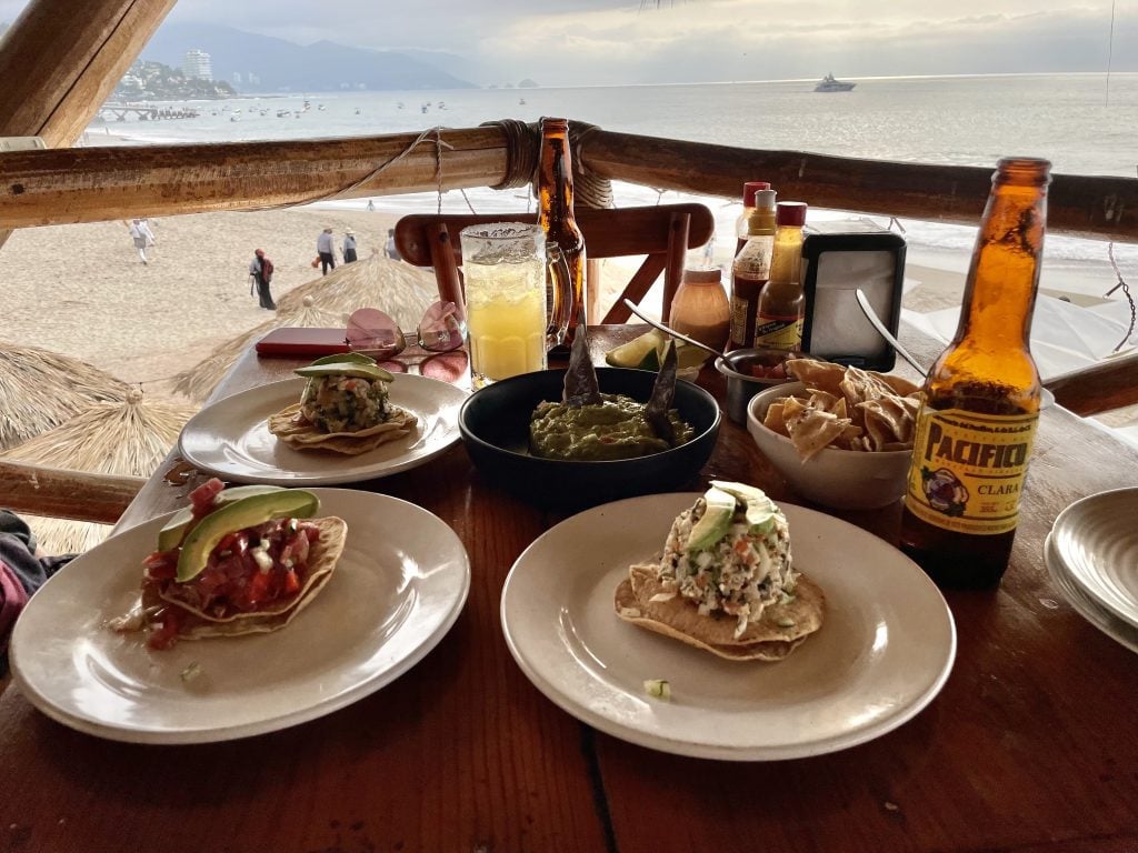 A restaurant table overlooking the beach. On the table are three tostadas and two bottles of beer.