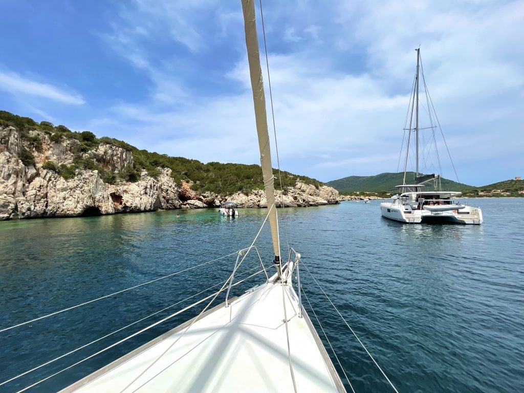 The front of a boat pointed out over a calm blue-green harbor surrounded by cliffs and another sailboat.