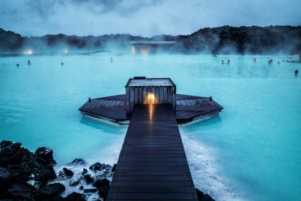 A wooden walkway leading to a hexagonal platform on top of the milky blue waters of the blue Lagoon, enshrouded in shadows at night.