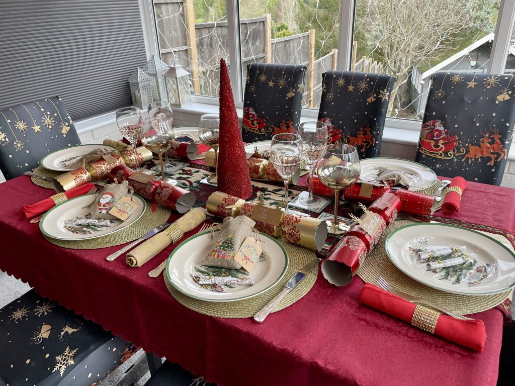 A table set for Christmas with a red tablecloth, red and gold napkins, little christmas trees, Christmas crackers, and chair covers with Santa and his reindeer on them.