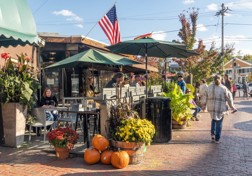 People walking past an outdoor cafe decorated with pumpkins and chrysanthemums.