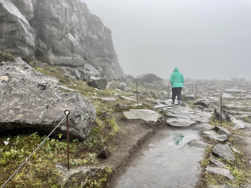 A girl in a turquoise coat walking on a rocky path in a foggy, rainy landscape.
