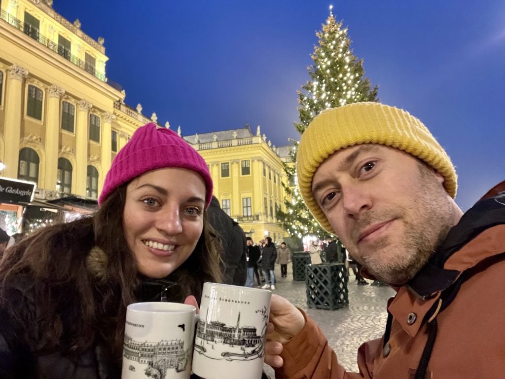 Kate and Charlie toasting gluhwein mugs with hats on in front of a bright yellow palace with a Christmas tree.