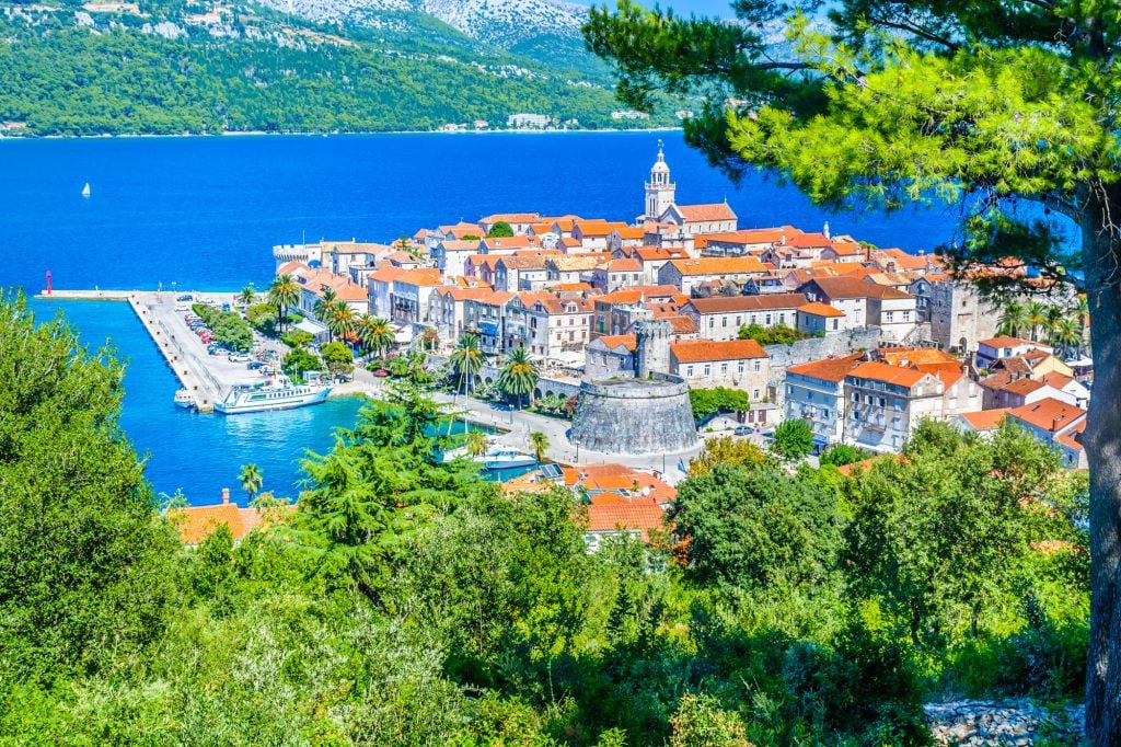 An aerial photo of Korcula town, a peninsula topped with orange-roofed stone buildings, surrounded by water and pine trees.