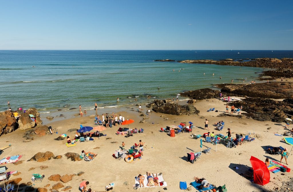 People relaxing on a long rocky beach leading to teal water.