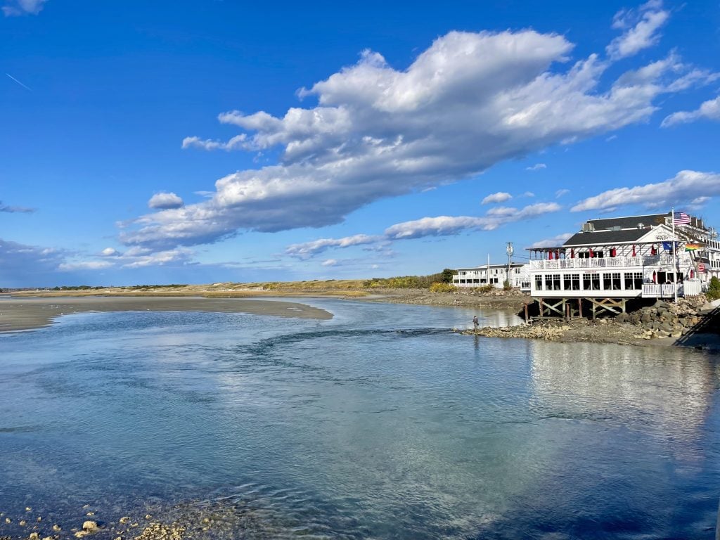 A big white wood building perched on a beach with shallow water flooding in front.