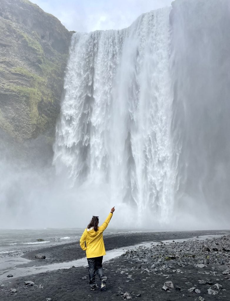 Kate standing facing a waterfall in a yellow rain jacket, holding her hand up in a peace sign.