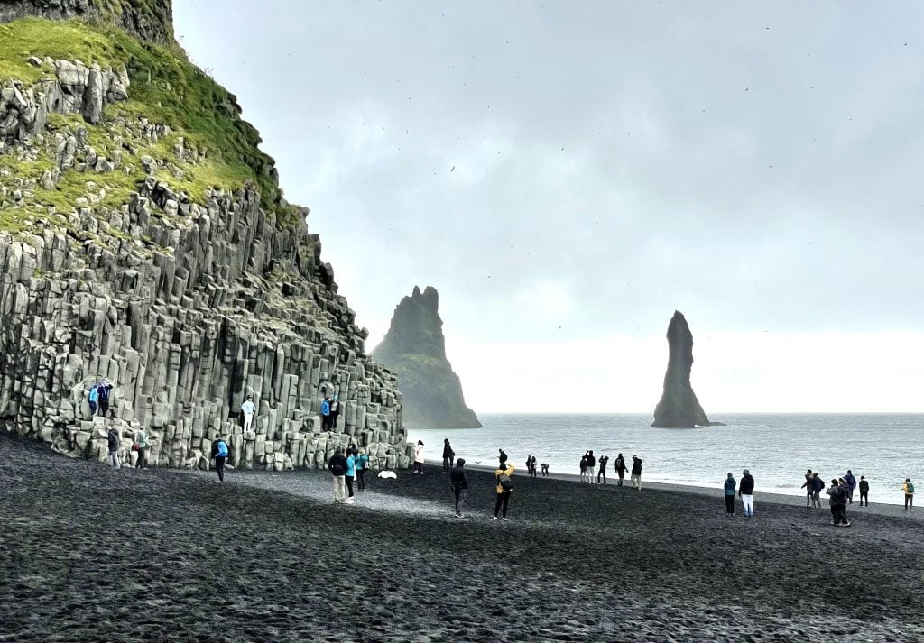 A black sand beach with lots of people standing dangerously close to the water.