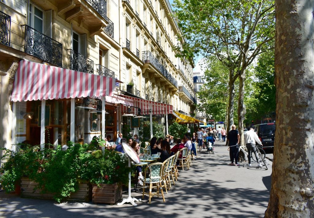People sitting at outdoor cafes on a leafy street in Paris.