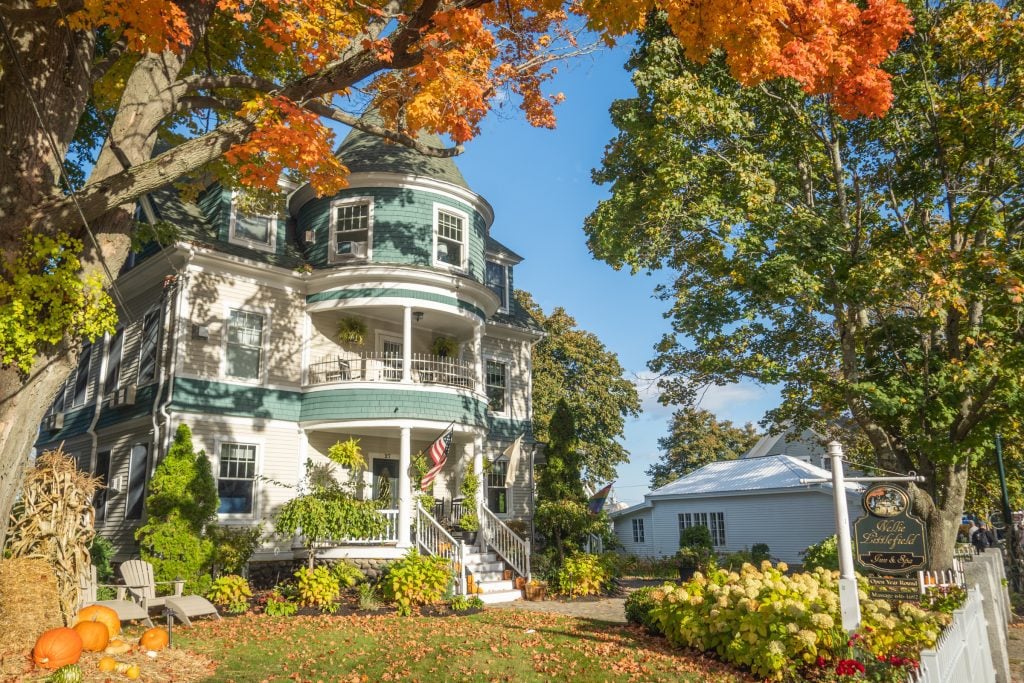 A victorian house with a turret underneath a big oak tree with bright orange leaves.