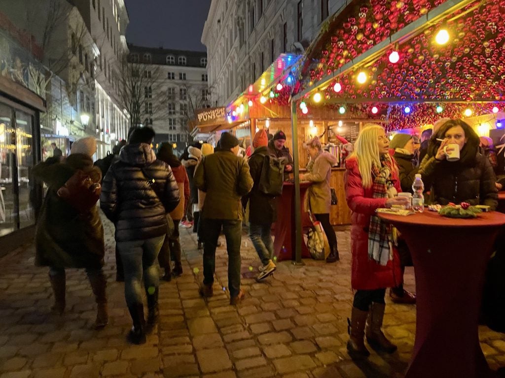 People strolling outside a pop-up bar in a market covered with Christmas lights.