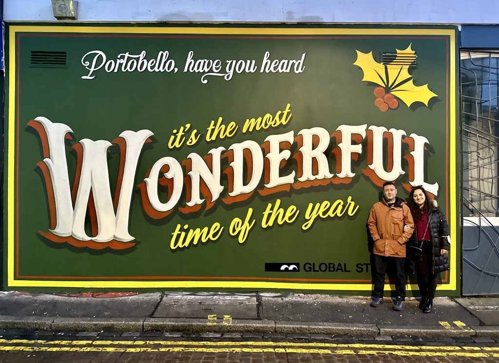 Kate and Charlie standing outside in coats in front of a sign reading "Portobello Road, it's the most wonderful time of the year"