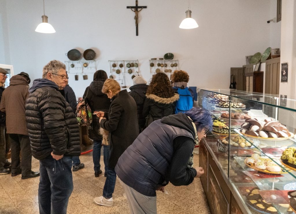 A simple bakery with pastries in cases and a prominent crucifix on the wall. An older woman leans down closely to look at the pastries.