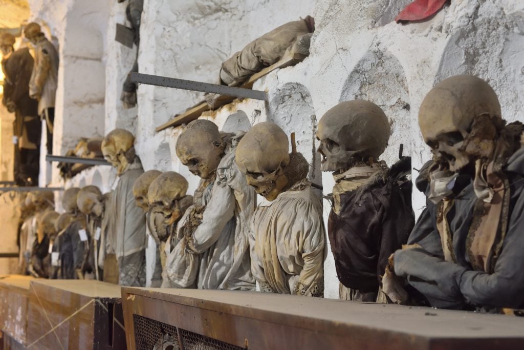 A row of creepy skeletons wearing clothes and lined up standing in the catacombs.