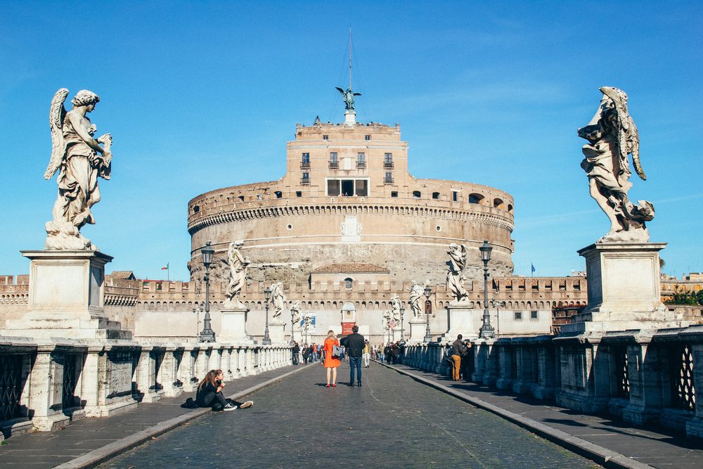 A couple standing in front of the Castel Sant'Angelo Italy Landmark on a clear day, with a blue sky in the background