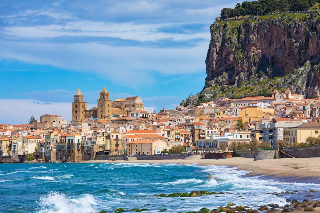 A gorgeous small town by the sea with sand-colored buildings, a few church towers, and rising cliffs behind it.