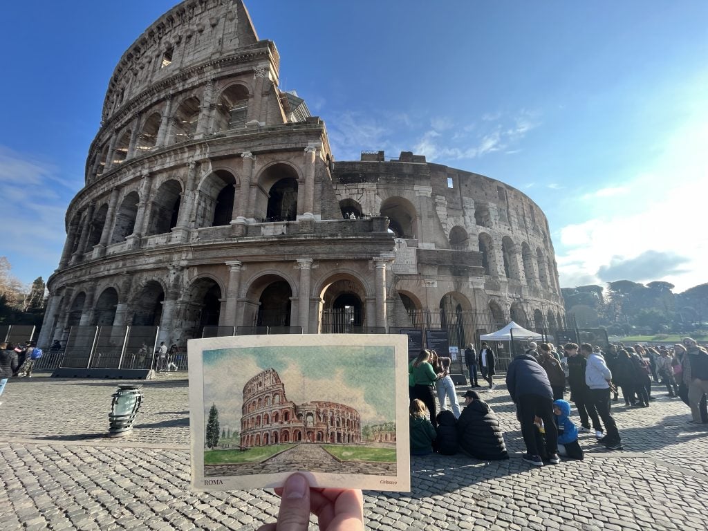 A hand holding a postcard of the Colosseum Italy landmark in front of the actual Colosseum