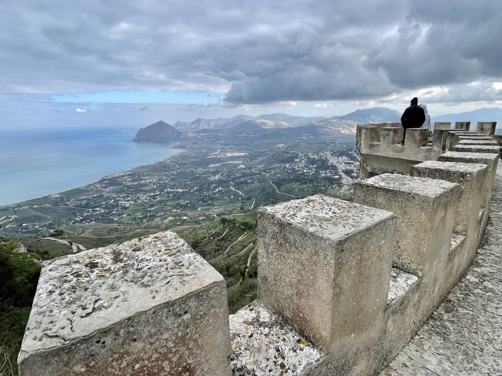 People standing on the edge of a stone castle in Erice, overlooking the coastline of Sicily in the distance.