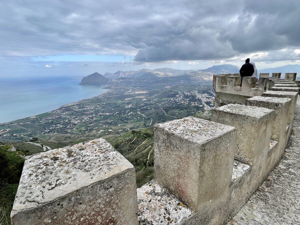 A couple standing on a stone fortress in Erice, looking over hills leading to the ocean.