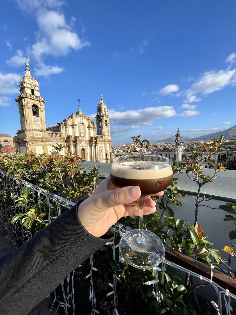A hand holding an espresso martini in front of a rooftop view of a white and yellow church on a piazza, blue sky behind it.