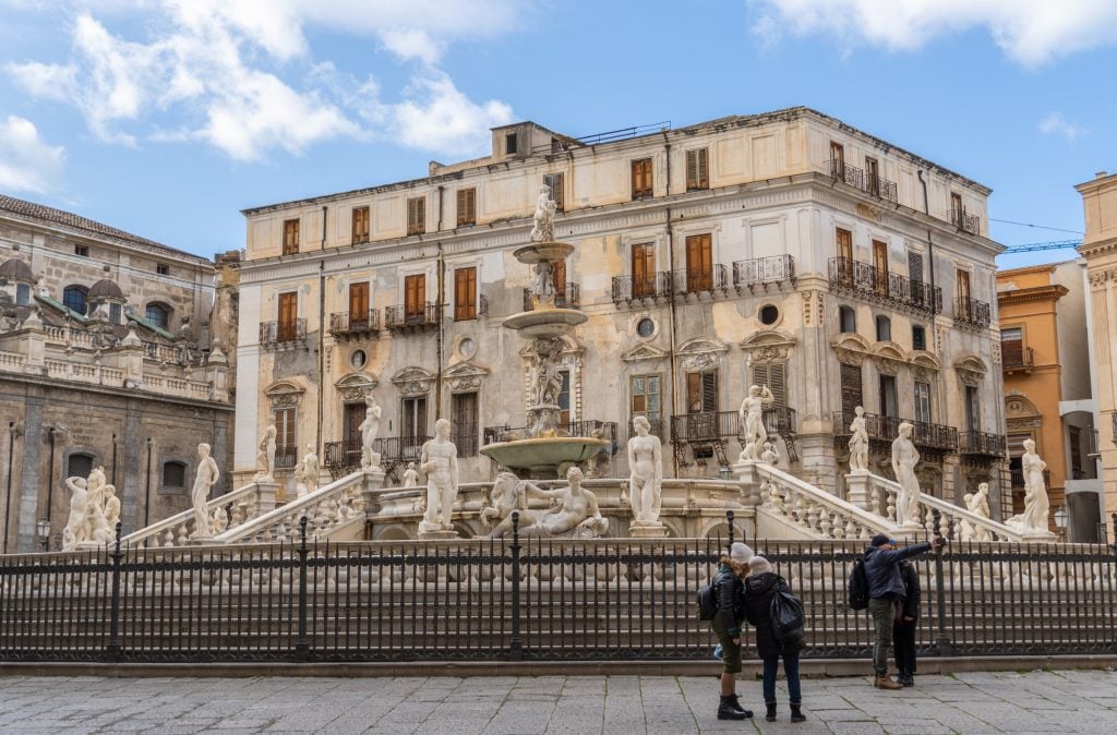 A Baroque fountain on an Italian piazza filled with lots of statues of muse-like women. In front of it some teenagers in winter coats take a selfie.