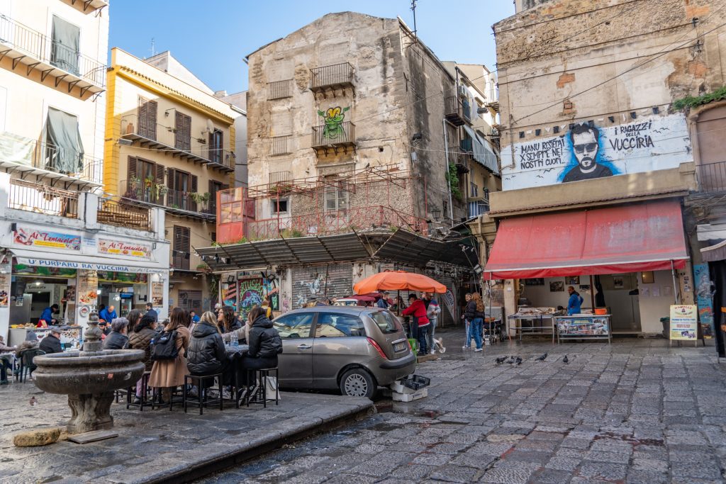 A market in Palermo, with lots of street art on the buildings and people eating at tables.
