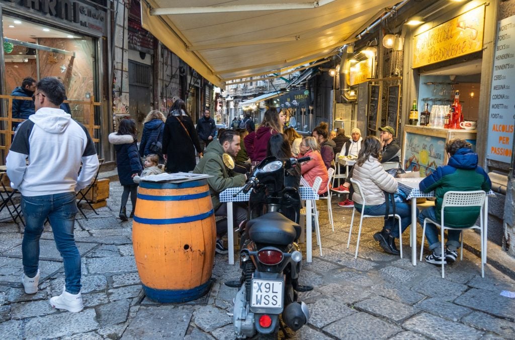 People sitting at street side restaurants, drinking spritzes and eating fried food.