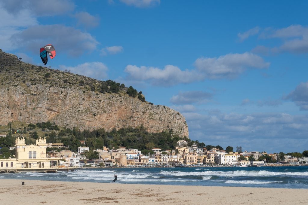 A beach in winter with big frothy waves and a kitesurfer surfing across them. Pastel-colored square beach buildings in the background.