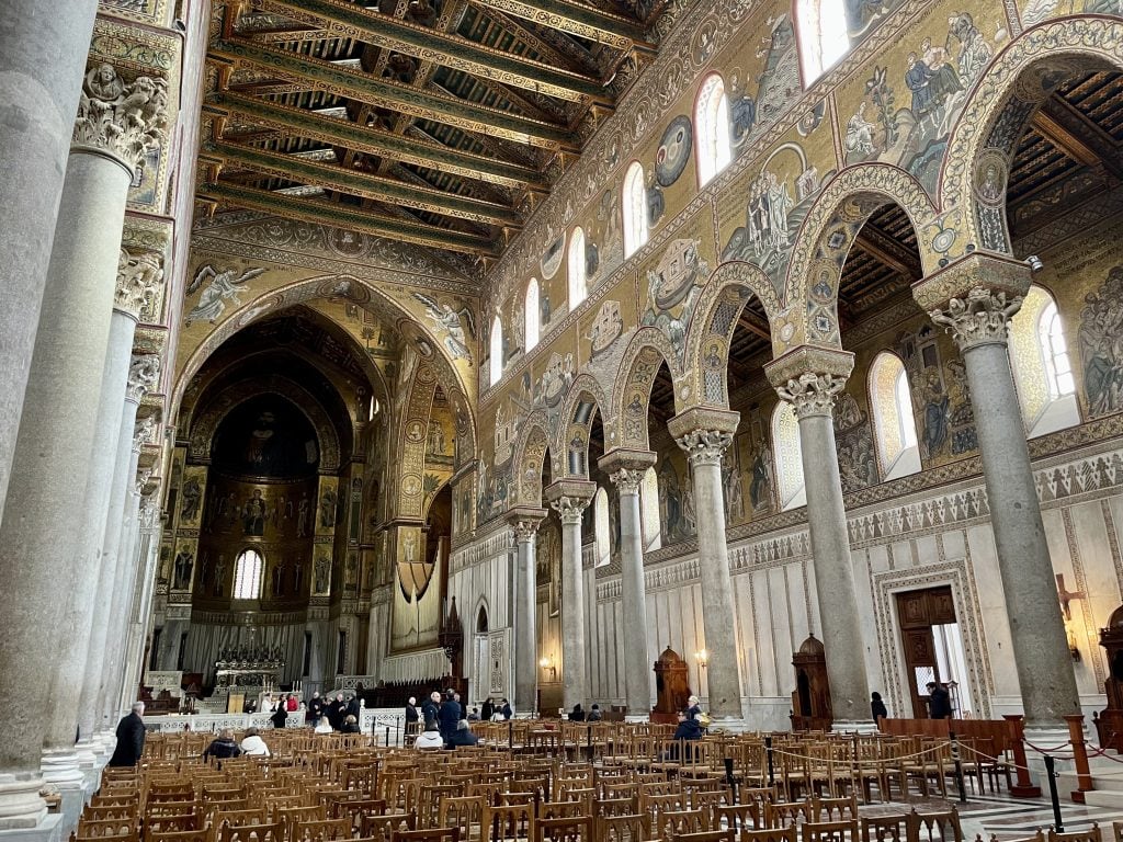 A church with tall columns along the aisles, a wooden ceilings, and gold mosaics on all the walls.