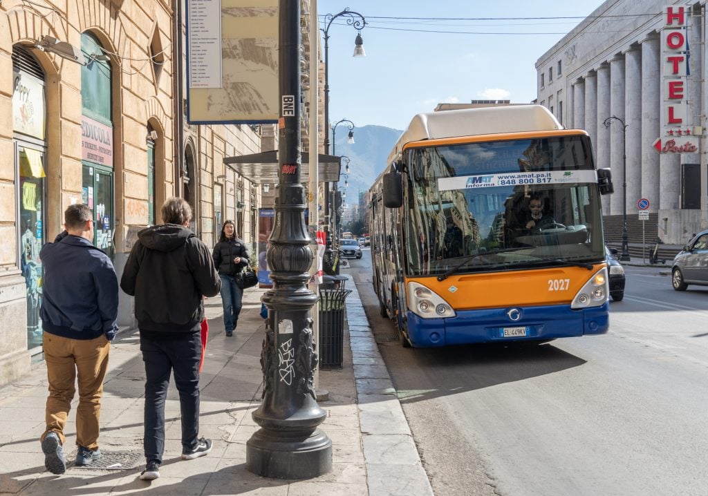 People walking on the sidewalk in Palermo as a bus drives by.