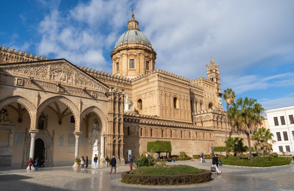 A sand-colored cathedral topped with a blue-green dome, with lots of crenellation on the outside and three big porticoes on the side.