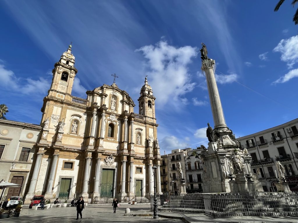 A Baroque Sicilian church with an obelisk in front underneath a blue sky.