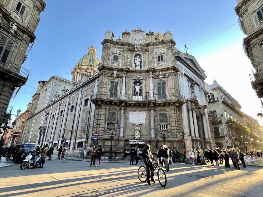 A person on a bicycle standing in front of a magnificent street corner filled with statues and a fountain.