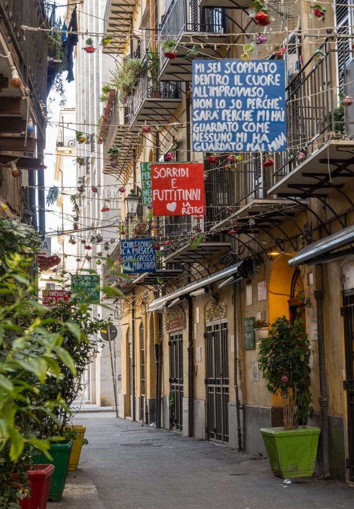 A narrow street in Palermo with flags and hand-lettered signs hanging between the buildings.